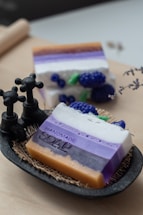 A decorative setup featuring handmade soap with layers of various colors, including orange, purple, and white, displayed in a small dish with a textured mat beneath. The soap includes a decorative element resembling blueberries nestled among the layers. Two antique-style black faucets are positioned near the soap, adding a rustic touch.
