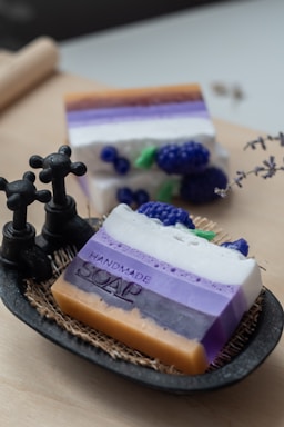 A decorative setup featuring handmade soap with layers of various colors, including orange, purple, and white, displayed in a small dish with a textured mat beneath. The soap includes a decorative element resembling blueberries nestled among the layers. Two antique-style black faucets are positioned near the soap, adding a rustic touch.