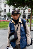 A person wearing a blue vest and cap is standing outdoors in a park. The vest has a logo and 'Municipalidad Metropolitana de Lima' written on it. The individual also wears a black neck scarf and gloves, carrying a camera bag or similar pouch. Behind them, there are green trees, a red flowerbed, and white colonial-style buildings.