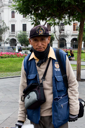 A person wearing a blue vest and cap is standing outdoors in a park. The vest has a logo and 'Municipalidad Metropolitana de Lima' written on it. The individual also wears a black neck scarf and gloves, carrying a camera bag or similar pouch. Behind them, there are green trees, a red flowerbed, and white colonial-style buildings.
