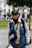 A person wearing a blue vest and cap is standing outdoors in a park. The vest has a logo and 'Municipalidad Metropolitana de Lima' written on it. The individual also wears a black neck scarf and gloves, carrying a camera bag or similar pouch. Behind them, there are green trees, a red flowerbed, and white colonial-style buildings.