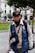 A person wearing a blue vest and cap is standing outdoors in a park. The vest has a logo and 'Municipalidad Metropolitana de Lima' written on it. The individual also wears a black neck scarf and gloves, carrying a camera bag or similar pouch. Behind them, there are green trees, a red flowerbed, and white colonial-style buildings.