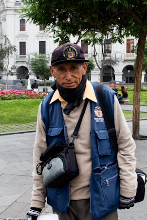 A person wearing a blue vest and cap is standing outdoors in a park. The vest has a logo and 'Municipalidad Metropolitana de Lima' written on it. The individual also wears a black neck scarf and gloves, carrying a camera bag or similar pouch. Behind them, there are green trees, a red flowerbed, and white colonial-style buildings.