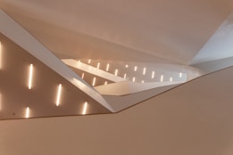 The interior features a modern architectural design with intersecting angular lines and surfaces. Soft, ambient lighting is provided by a row of vertical light fixtures along the walls. The space is largely composed of smooth, white surfaces, with light creating patterns of shadows.