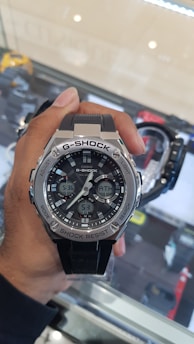 A hand is holding a large, rugged G-Shock wristwatch with a metallic bezel. The watch features multiple digital displays indicating the time and other functionalities. The background shows a glass display case with another watch and some reflections.