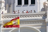 A group of people stand on a balcony of an ornate building with architectural detailing. A large Spanish flag is prominently displayed in the foreground, waving against a clear blue sky.