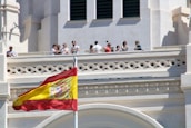A group of people stand on a balcony of an ornate building with architectural detailing. A large Spanish flag is prominently displayed in the foreground, waving against a clear blue sky.
