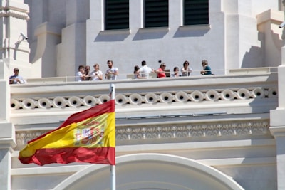 A group of people stand on a balcony of an ornate building with architectural detailing. A large Spanish flag is prominently displayed in the foreground, waving against a clear blue sky.