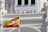 A group of people stand on a balcony of an ornate building with architectural detailing. A large Spanish flag is prominently displayed in the foreground, waving against a clear blue sky.