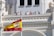 A group of people stand on a balcony of an ornate building with architectural detailing. A large Spanish flag is prominently displayed in the foreground, waving against a clear blue sky.