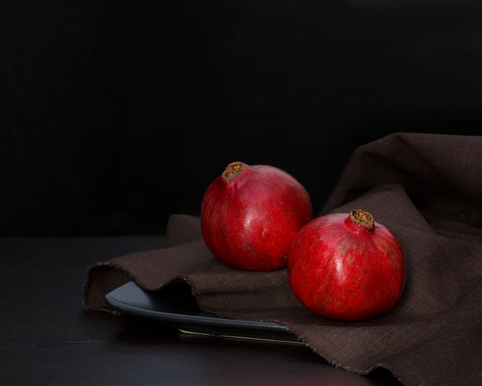 Two ripe red pomegranates rest on a black plate on a brown tablecloth