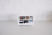 A smart display device sits on a white kitchen counter against a backdrop of white tiled walls. The screen displays various interactive options such as Find food recipes, Play videos, Manage photos, and Check news, each represented by images.