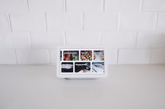 A smart display device sits on a white kitchen counter against a backdrop of white tiled walls. The screen displays various interactive options such as Find food recipes, Play videos, Manage photos, and Check news, each represented by images.