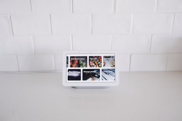 A smart display device sits on a white kitchen counter against a backdrop of white tiled walls. The screen displays various interactive options such as Find food recipes, Play videos, Manage photos, and Check news, each represented by images.