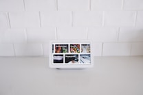 A smart display device sits on a white kitchen counter against a backdrop of white tiled walls. The screen displays various interactive options such as Find food recipes, Play videos, Manage photos, and Check news, each represented by images.