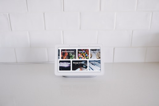 A smart display device sits on a white kitchen counter against a backdrop of white tiled walls. The screen displays various interactive options such as Find food recipes, Play videos, Manage photos, and Check news, each represented by images.