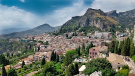 A picturesque town nestled in a valley surrounded by rugged mountains. The town is densely populated with charming buildings topped with terracotta roofs, interspersed with lush greenery and tall cypress trees. The landscape features dramatic rocky cliffs in the background, with a partly cloudy sky above.