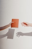 A hand on the left is holding a red notebook with the word '2020' embossed on it, while a hand on the right is reaching out as if to receive it. The neutral background casts shadows of both hands and the notebook, creating a simple and artistic composition.