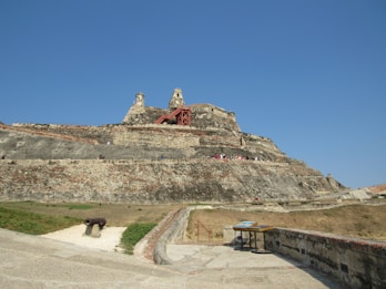 A large, historic fortress with multiple tiers and stone walls set against a clear blue sky. The structure has red staircases and scattered vegetation around its base. A historical cannon is positioned on the ground nearby, with a spacious paved area leading up to the fortress.