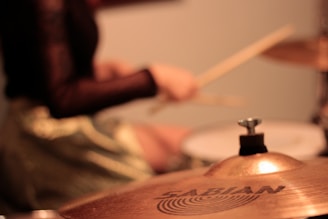 A close-up view of a drum set, focusing on a cymbal with the brand name Sabian visible. In the background, a person is playing drums with drumsticks in hand, creating a dynamic and musical atmosphere.