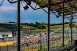 A racetrack scene with a large crowd gathered along the side of the track, possibly in anticipation of an event. Rows of empty spectator seats are visible in the foreground under a metal roof structure. In the background, the track is lined with people, indicating an organized or official event. The surrounding landscape includes green hills and trees under a cloudy blue sky.
