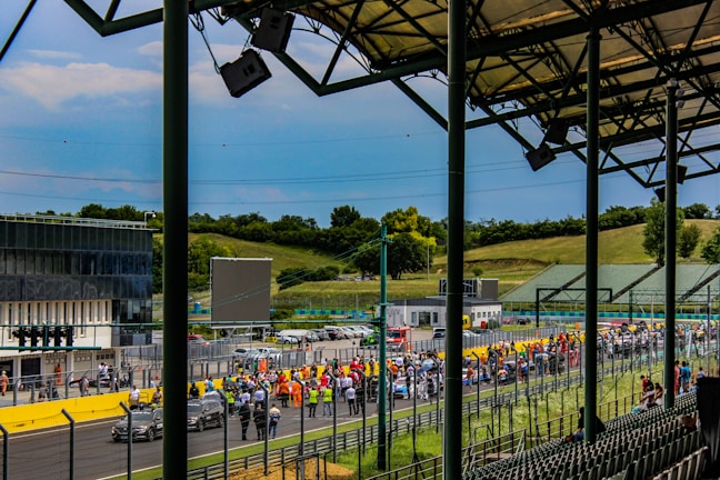 A racetrack scene with a large crowd gathered along the side of the track, possibly in anticipation of an event. Rows of empty spectator seats are visible in the foreground under a metal roof structure. In the background, the track is lined with people, indicating an organized or official event. The surrounding landscape includes green hills and trees under a cloudy blue sky.