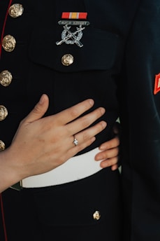 A person is wearing a formal military uniform adorned with gold buttons and badges. Another individual’s hand, wearing a diamond ring, is gently placed on the arm of the uniformed person, conveying a sense of intimacy and connection.