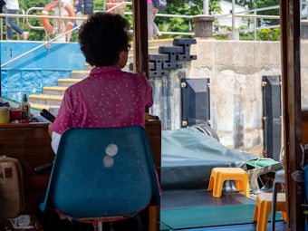 An individual sits on a blue chair inside a wooden structure, facing towards a dock area with steps and a blue wall. There are orange stools on a platform outside, and various boating equipment is visible. Greenery can be seen in the background, indicating a riverside or lakeside setting.