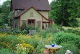 A quaint two-story house with a sloped roof nestled in a lush garden filled with a variety of colorful flowers and greenery. A wooden bench sits in front of the house, surrounded by climbing plants. A birdbath with a blue glass ornament is prominently placed in the foreground, adding a whimsical touch to the natural scenery. Tall trees and verdant plants envelop the house, creating a serene, secluded atmosphere.