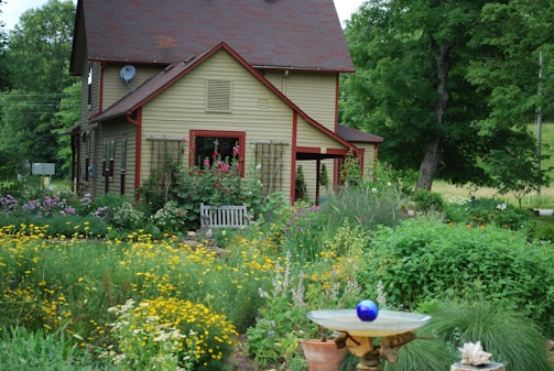 A quaint two-story house with a sloped roof nestled in a lush garden filled with a variety of colorful flowers and greenery. A wooden bench sits in front of the house, surrounded by climbing plants. A birdbath with a blue glass ornament is prominently placed in the foreground, adding a whimsical touch to the natural scenery. Tall trees and verdant plants envelop the house, creating a serene, secluded atmosphere.