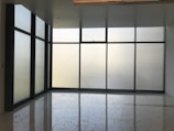 Large floor-to-ceiling glass windows are framed in dark metal, creating a modern and minimalistic design. The frosted glass provides a blurred view of the outside, allowing softened light to filter into the room. The floor is polished and reflects the light softly, adding to the clean and uncluttered aesthetic.