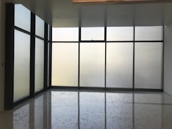 Large floor-to-ceiling glass windows are framed in dark metal, creating a modern and minimalistic design. The frosted glass provides a blurred view of the outside, allowing softened light to filter into the room. The floor is polished and reflects the light softly, adding to the clean and uncluttered aesthetic.