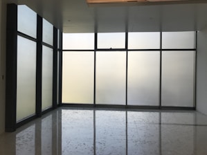 Large floor-to-ceiling glass windows are framed in dark metal, creating a modern and minimalistic design. The frosted glass provides a blurred view of the outside, allowing softened light to filter into the room. The floor is polished and reflects the light softly, adding to the clean and uncluttered aesthetic.