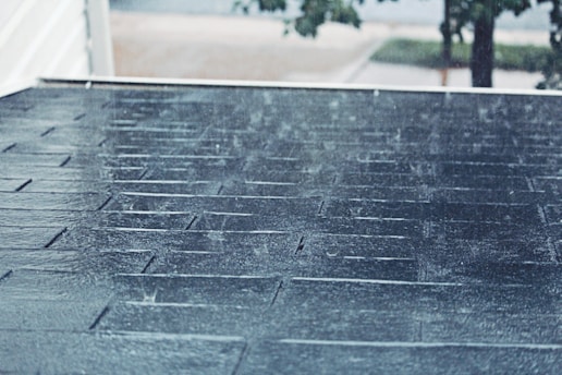 Rain is falling on a sloped shingle roof creating a wet and shiny surface. The background is slightly blurred, showing hints of trees and a street, suggesting a suburban environment. The rain creates a serene yet somber atmosphere.