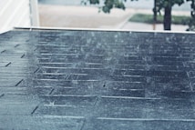 Rain is falling on a sloped shingle roof creating a wet and shiny surface. The background is slightly blurred, showing hints of trees and a street, suggesting a suburban environment. The rain creates a serene yet somber atmosphere.