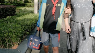 A child wearing a blue and green jacket and a black shirt with the letters 'CR7' is seen walking beside an adult who is wearing a sheer black dress. The child holds a blue bag decorated with cartoon images and British-themed icons. They are surrounded by green bushes and are walking on a paved path, typical of a park setting.