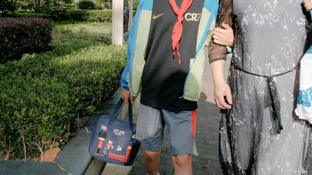 A child wearing a blue and green jacket and a black shirt with the letters 'CR7' is seen walking beside an adult who is wearing a sheer black dress. The child holds a blue bag decorated with cartoon images and British-themed icons. They are surrounded by green bushes and are walking on a paved path, typical of a park setting.