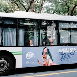 A bus traveling along a street with passengers visible through the windows. An advertisement is prominently displayed on the side of the bus featuring a smiling woman holding beauty products. The bus is parked against a backdrop of lush green trees.