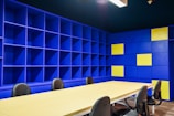 A modern office space with a large beige conference table surrounded by black chairs. The walls are covered with blue shelving units, some sections of which are highlighted with yellow panels. The lighting is bright, giving the room an open and vibrant feel.