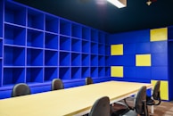 A modern office space with a large beige conference table surrounded by black chairs. The walls are covered with blue shelving units, some sections of which are highlighted with yellow panels. The lighting is bright, giving the room an open and vibrant feel.