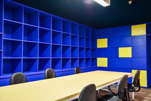 A modern office space with a large beige conference table surrounded by black chairs. The walls are covered with blue shelving units, some sections of which are highlighted with yellow panels. The lighting is bright, giving the room an open and vibrant feel.