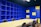 A modern office space with a large beige conference table surrounded by black chairs. The walls are covered with blue shelving units, some sections of which are highlighted with yellow panels. The lighting is bright, giving the room an open and vibrant feel.