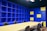 A modern office space with a large beige conference table surrounded by black chairs. The walls are covered with blue shelving units, some sections of which are highlighted with yellow panels. The lighting is bright, giving the room an open and vibrant feel.