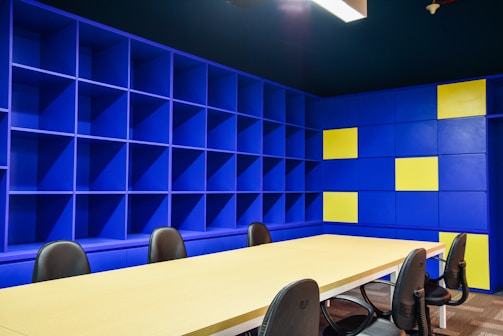 A modern office space with a large beige conference table surrounded by black chairs. The walls are covered with blue shelving units, some sections of which are highlighted with yellow panels. The lighting is bright, giving the room an open and vibrant feel.