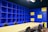 A modern office space with a large beige conference table surrounded by black chairs. The walls are covered with blue shelving units, some sections of which are highlighted with yellow panels. The lighting is bright, giving the room an open and vibrant feel.