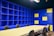 A modern office space with a large beige conference table surrounded by black chairs. The walls are covered with blue shelving units, some sections of which are highlighted with yellow panels. The lighting is bright, giving the room an open and vibrant feel.
