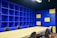 A modern office space with a large beige conference table surrounded by black chairs. The walls are covered with blue shelving units, some sections of which are highlighted with yellow panels. The lighting is bright, giving the room an open and vibrant feel.