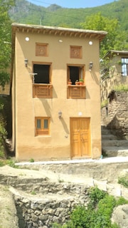 A traditional two-story adobe house with wooden windows and door, adorned with lattice designs. The structure is set against a backdrop of lush green hills, with foliage surrounding the area. Clay pots with plants embellish the window sills, and a stone pathway leads to the entrance.