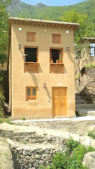 A traditional two-story adobe house with wooden windows and door, adorned with lattice designs. The structure is set against a backdrop of lush green hills, with foliage surrounding the area. Clay pots with plants embellish the window sills, and a stone pathway leads to the entrance.