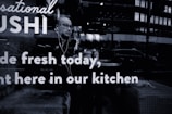 A person wearing glasses and earphones is seated at a table inside a sushi restaurant. The person appears to be looking outside through the window. Reflections from the street outside, including a few vehicles and buildings, are visible on the glass. The words 'Sushi made fresh today, right here in our kitchen' are prominently displayed on the window glass.
